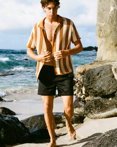 A man in Dandy Del Mar’s The Mallorca Swim-Walk Shorts – Onyx, featuring adjustable side fasteners, walks barefoot on a rocky beach as waves break in the background.