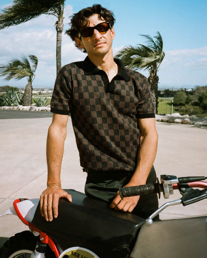 A person in The Sebastian Knit Polo - Onyx from Dandy Del Mar and sunglasses stands outdoors, hand on a motorcycle, with wind-swayed trees. The scene exudes a relaxed vibe akin to wearing a summer sweater in cool breezes or experiencing the refined touch of an Italian knit.