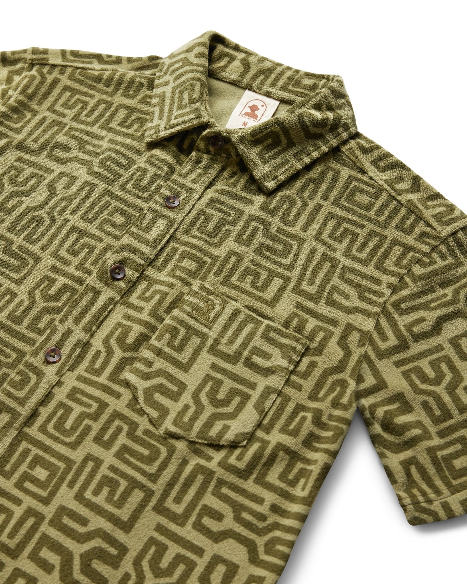 The Tropez Terry Cloth Shirt - Berdea Tayana Print by Dandy Del Mar is a short-sleeve button-up with a green geometric maze pattern, chest pocket, and an inner collar brand tag, inspired by the Côte d'Azur’s relaxed vibe.